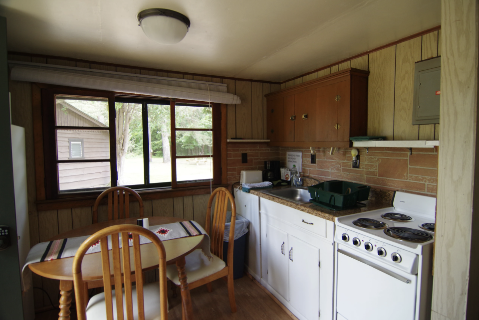 A quaint, cozy cottage kitchen with small dining table in front of windows with views of woods.