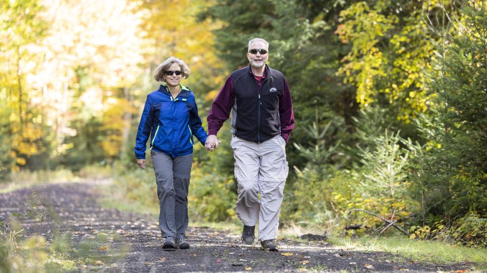 An older couple in hiking clothes walk along a path, holding hands and smiling.