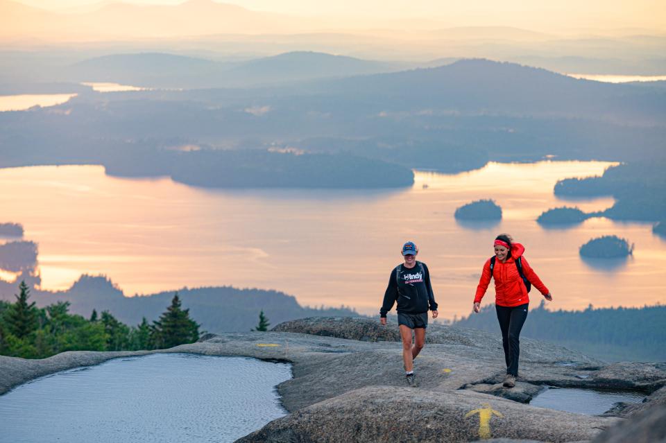 Two people walk along an open rocky summit with a large lake behind them