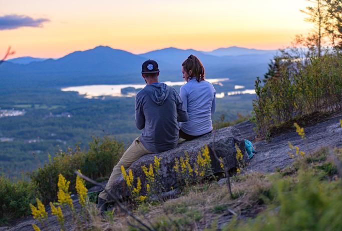 A man and woman sit on a mountain summit in the warm colors of a spring sunset.