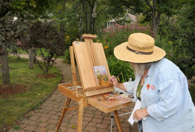 A person in a straw hat paints at an easel in a park, surrounded by greenery.