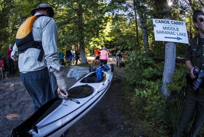 Spectators watch groups of Adirondack 90-Miler participants leaving the water and carrying their vessels towards Middle Saranac Lake on a dirt trail