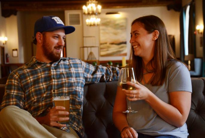 A couple smiles at each other while enjoying a beer and wine as they sit on a couch