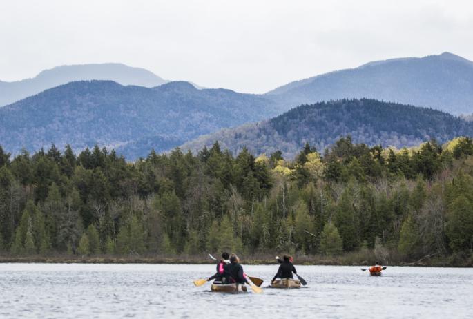 Two canoes and a kayak paddle away from the camera and towards the backdrop of the Adirondack mountains
