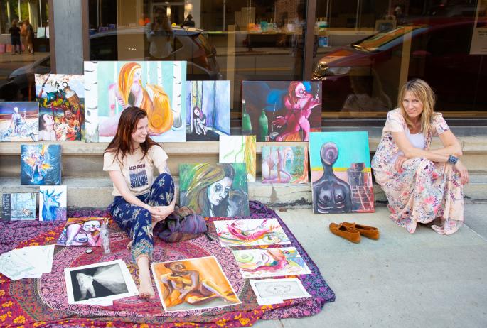 Two female artists sit on the sidewalk with their paintings during Third Thursday Art Walk in Saranac Lake