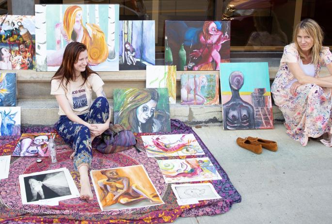 Two women display visual arts on a sidewalk.