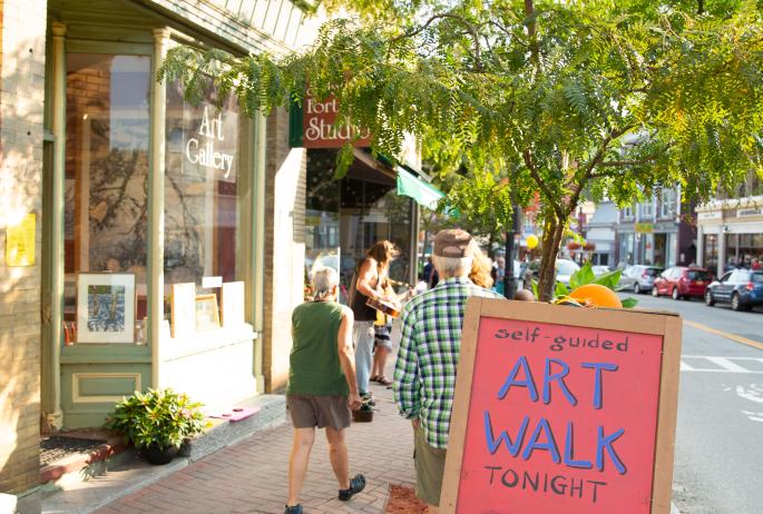 A colorful Art Walk sign stands before a busy sidewalk with artists and visitors.