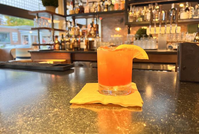 A pink cocktail with an orange slice at a bar.