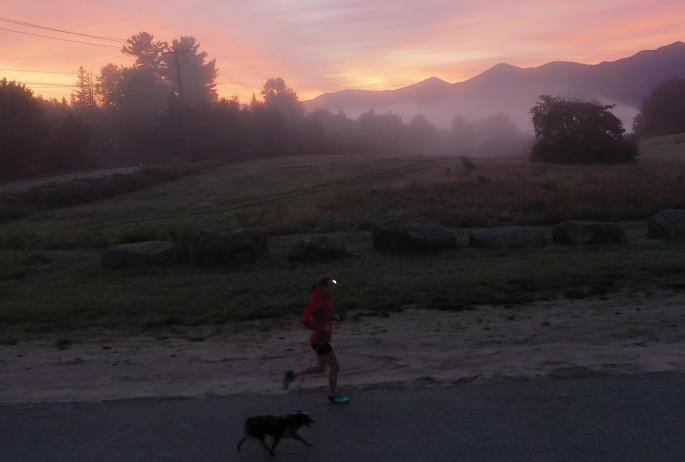 An athlete runs up a road with her dog as the sun rises among the mountains behind them