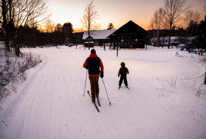 An adult and child cross-country ski at sunset on groomed trails.