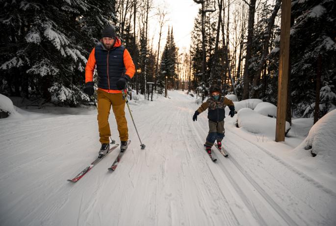 An adult and child cross-country ski at sunset on groomed trails through a forest.