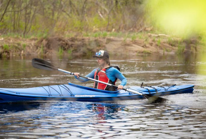 A woman in a blue kayak paddles down a river amid bright green spring vegetation.