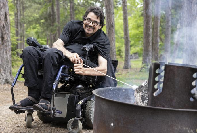 A man in a motorized wheelchair toasts a marshmallow over a campfire