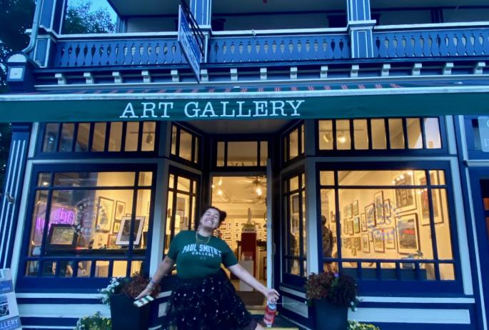 The exterior of a charming, brightly lit art gallery with awning over the door. A college student poses happily outside the door.
