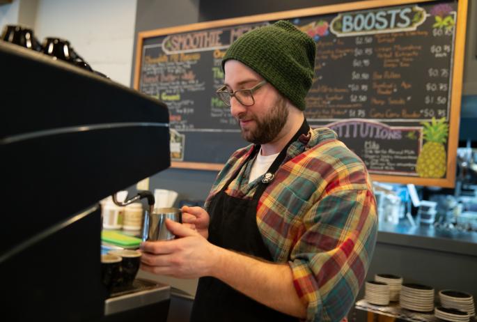 A barista wearing a flannel and green winter hat creates a drink with a menu behind him.