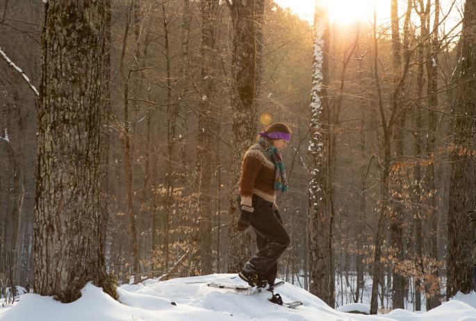 A woman snowshoes through a forest illuminated by morning sunlight