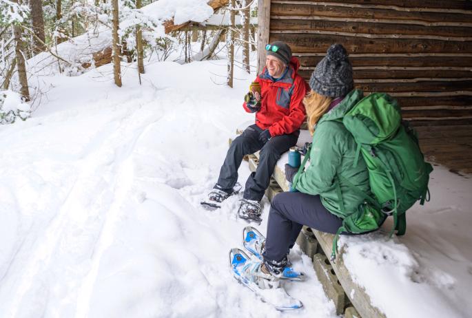 Two friends take a break from snowshoeing and enjoy a drink of water in a lean to in a snow covered forest