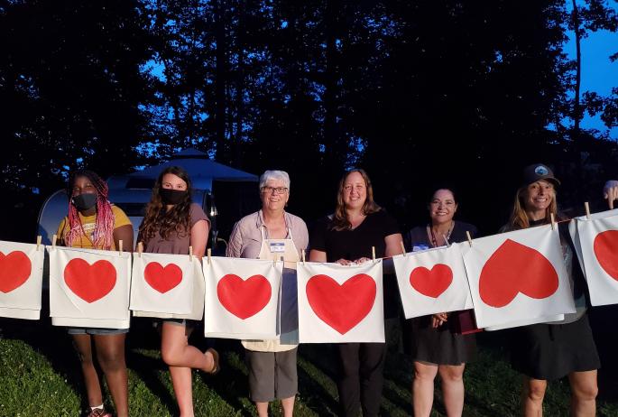 Six women stand behind a clothesline that holds a number of red hearts printed on white fabric.