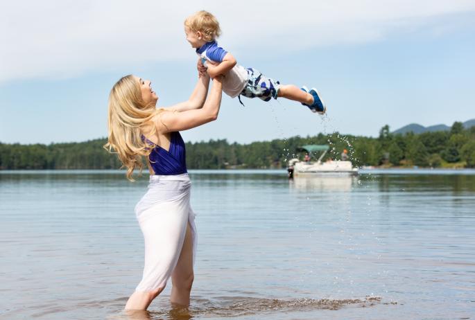 A blonde woman in a bathing suit lifts a smiling toddler out of a sparkling lake. A boat and trees are on the shore beyond.