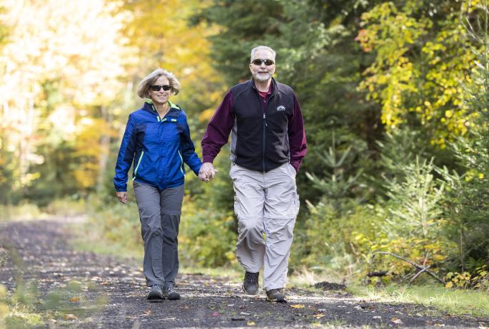 An older couple in hiking clothes walk along a path, holding hands and smiling.