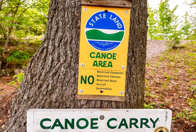 A state land sign nailed to a tree marking the boundary into the Canoe Area, with a canoe carry sign below it