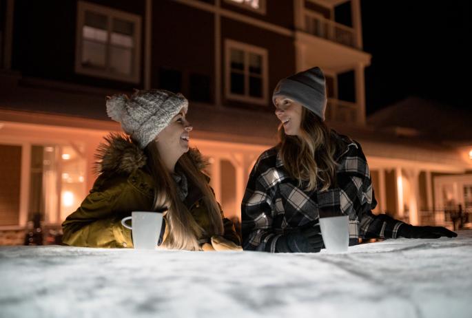 Two women laugh over hot cocoa in front of a hotel at night.