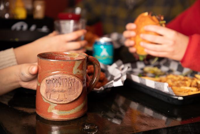 A close up of a person holding a burger while another holds a mug with a rustic "Grizle T's" logo on it.