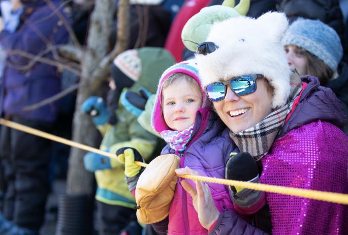 A woman wears sunglasses and a polar bear hat as she smiles with a young child.
