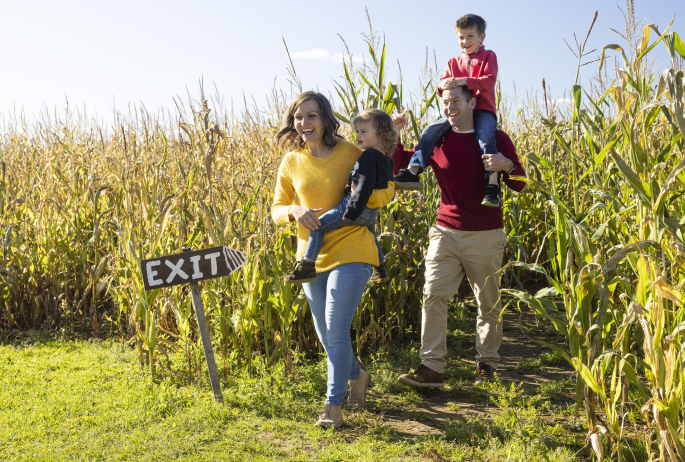 A man and woman carry their two young children out of a corn maze.