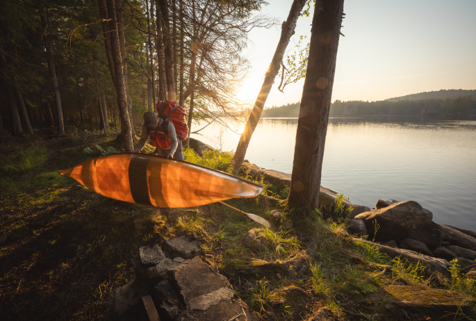 A person carrying a canoe and gear to a pond.