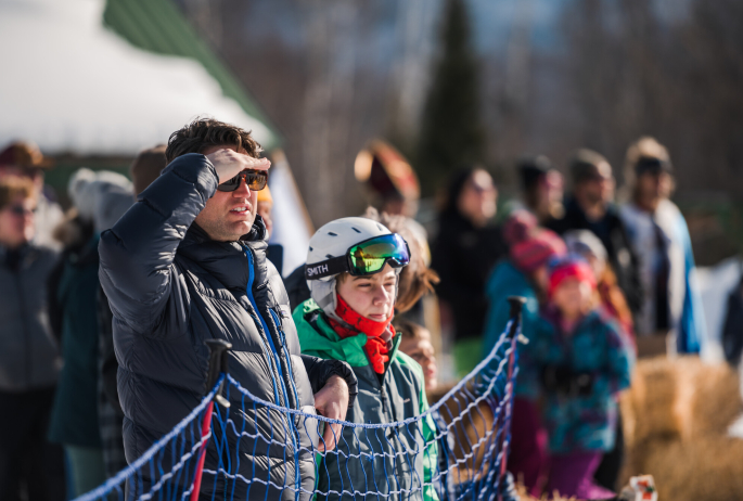 A child in a ski helmet and goggles stands with his dad while watching a winter outdoor event.