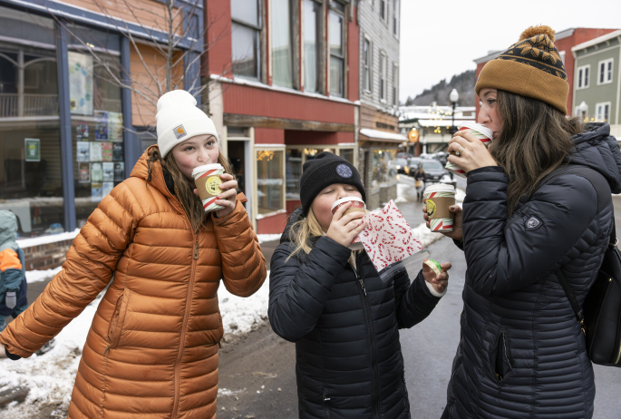 Three women smile and sip hot cocoa on a winter street.