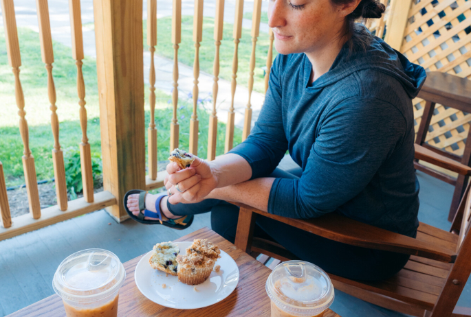 A woman sits in a rocking chair with a muffin and iced coffee on a small table next to her.