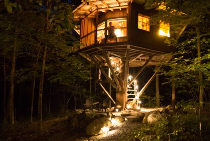 A man stands at the top of a treehouse at night.