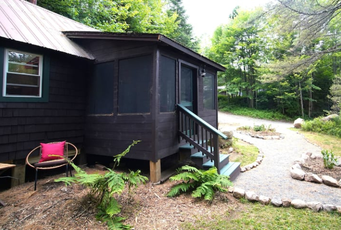 A dark brown cabin sits at the base of a driveway in the woods.