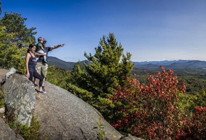A couple holding a toddler points off into the distance from the top of Baker Mountain with some of the trees closest to them starting to turn red
