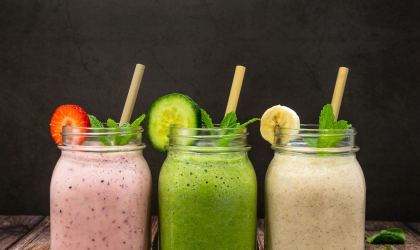 Three colorful mason jars of fresh cold pressed juice on a table surrounded by mint leaves and blueberries.