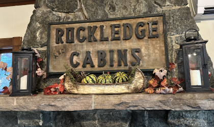A carved sign reading 'Rockledge and Cabins' over a stone mantle with birch bark boat filled with festive squash and tall candles.